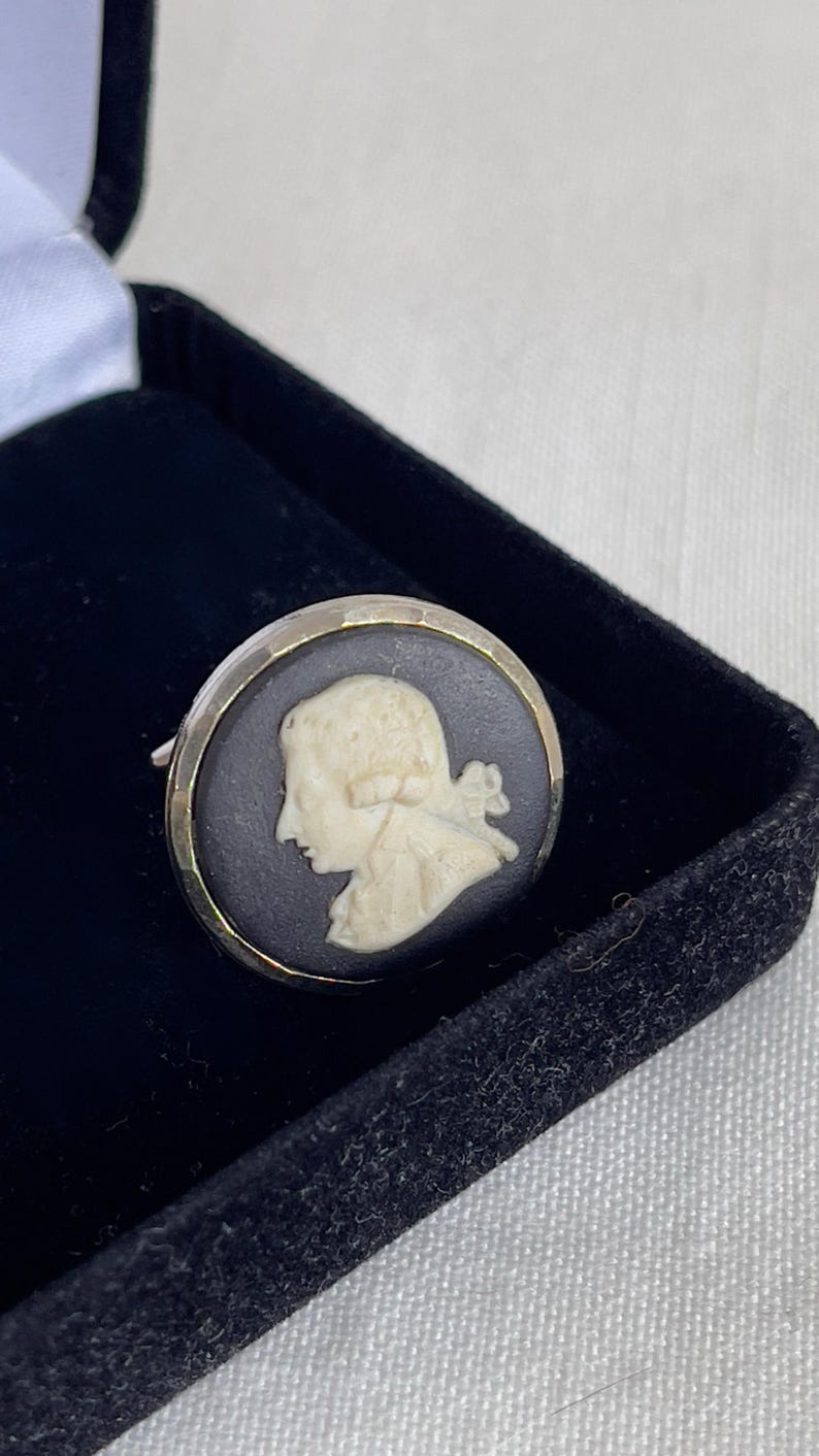 May include: A vintage cameo ring featuring a cream-colored profile set against a black background. The ring has a silver-colored metal band and is displayed in a black velvet box. The cameo depicts a classical figure.