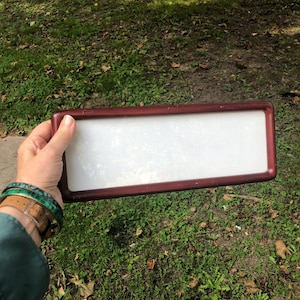 May include: A rectangular frame with a dark reddish-brown finish and a smooth, opaque white center panel. The frame has rounded corners and a simple, clean design, providing a neutral surface that is held horizontally against a natural outdoor setting.