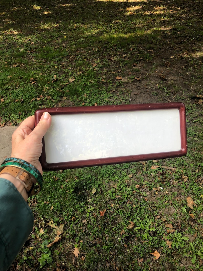May include: A rectangular frame with a dark reddish-brown finish and a smooth, opaque white center panel. The frame has rounded corners and a simple, clean design, providing a neutral surface that is held horizontally against a natural outdoor setting.