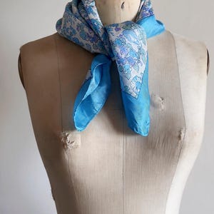 May include: A square scarf tied around the neck of a mannequin. The scarf features a central white panel with a repeating floral pattern in shades of light blue, periwinkle, and soft green, bordered by a solid bright blue edge on all sides.