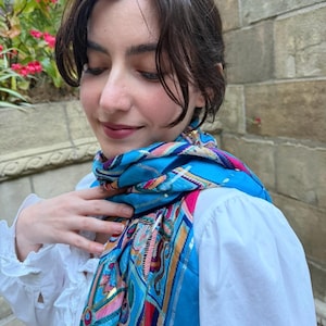 May include: A bright blue scarf featuring a multi-coloured geometric and abstract pattern in shades of pink, gold, and dark blue, worn over a white long-sleeved shirt with ruffled detailing along the front placket.