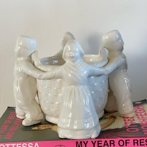 Puede incluir: Una maceta de cerámica blanca con un diseño en forma de cesta, rodeada por cuatro figuras con vestimenta tradicional. Las figuras se toman de la mano, creando una disposición circular alrededor de la maceta. La maceta está sobre un libro.