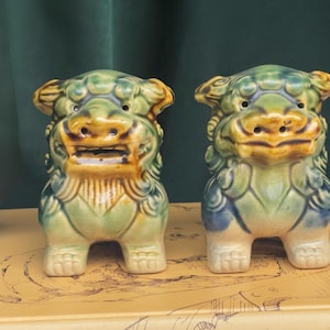 Może przedstawiać: Dwie ceramiczne figurki psów Foo. Posągi są zielone, niebieskie i żółte ze szczegółowymi detalami. Stoją na beżowej powierzchni z widocznymi słowami "CARD ON BREAD". Tło to ciemnozielona zasłona.
