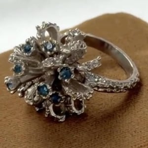 Puede incluir: Un anillo de tono plateado que presenta un grupo de bucles metálicos texturizados en forma de cinta dispuestos en una forma esférica. Varias piedras pequeñas, redondas y de color azul oscuro están engastadas en la intrincada estructura metálica.