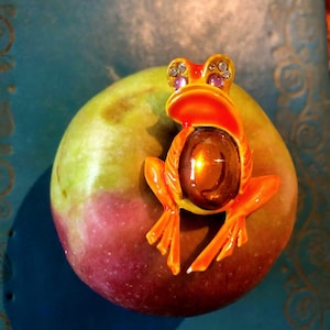 Peut inclure: Une figurine décorative en forme de grenouille orange et jaune avec des accents violets, posée sur une pomme verte et rouge. La grenouille a un grand ventre brun en forme de bijou. La pomme est sur une surface bleue.