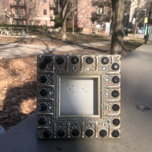 May include: A square picture frame with a metallic silver finish, featuring a decorative border embedded with alternating large black circular stones and smaller clear crystals. The frame has a central square opening for a photograph.