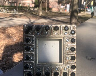 May include: A square picture frame with a metallic silver finish, featuring a decorative border embedded with alternating large black circular stones and smaller clear crystals. The frame has a central square opening for a photograph.