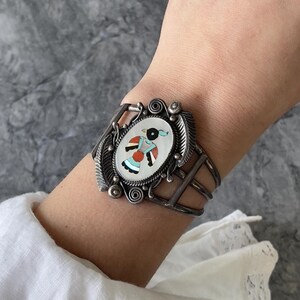 May include: A silver cuff bracelet featuring an oval inlay of a stylised bird figure. The inlay uses white, turquoise, red, and black stone segments. The silver frame is decorated with etched feather patterns, small metal spheres, and scrolls.