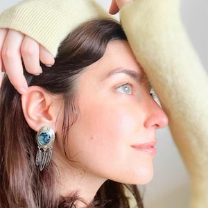 Peut inclure: Vue de profil d'une personne portant une boucle d'oreille en argent avec une pierre bleu-vert et des breloques en forme de plumes. La personne a les cheveux foncés et porte un pull jaune pâle. La boucle d'oreille est au centre.