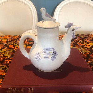 May include: A white ceramic teapot with a bird-shaped spout and a bird figurine on the lid. The teapot has a handle and is decorated with purple and yellow brushstrokes. The teapot is sitting on a red book.