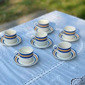 May include: A set of seven white ceramic teacups and saucers. Each cup and saucer features a design of blue and gold stripes. The cups have a handle and are placed on a white tablecloth with a floral design.