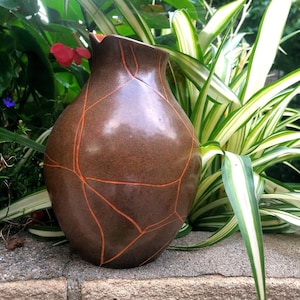 May include: A bulbous, dark brown ceramic vase with a smooth, glossy finish. The surface is decorated with a network of thin, bright orange lines that create a geometric, cracked pattern across the rounded body and the narrow, flared opening.