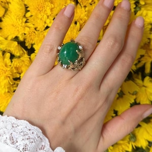 Op de afbeelding: Een ring met een grote, ovale, groenkleurige cabochon-geslepen steen in een textuurrijke, takachtige gouden band. De zetting bevat kleine, heldere, rondgeslepen accentstenen die zijn ingebed in het organische, twijgachtige metaalwerk rondom.
