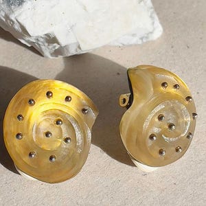 Könnte beinhalten: Ein Paar Clip-Ohrringe in einem spiralförmigen Muscheldesign. Die Ohrringe sind durchscheinend gelb mit silberfarbenen Nieten. Die Ohrringe haben einen Clipverschluss. Schmuck.