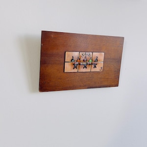 May include: A wooden wall art piece with a rectangular shape and a dark brown finish. It features a series of small, square tiles with a colourful illustration of people. The artwork is mounted on a white wall.