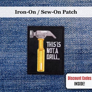 Puede incluir: Parche rectangular negro con plancha que presenta un martillo con cabeza plateada y mango amarillo. El texto "THIS IS NOT A DRILL" está debajo. Sobre un fondo de mezclilla azul.