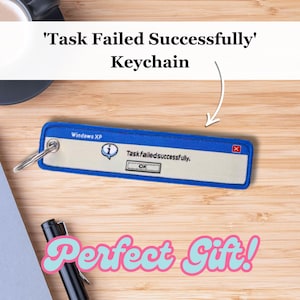 Może przedstawiać: Niebiesko-biały brelok w kształcie komunikatu o błędzie z systemu Windows XP. Komunikat brzmi "Zadanie nie powiodło się, zakończone sukcesem". Brelok leży na drewnianej powierzchni z długopisem i notesem.