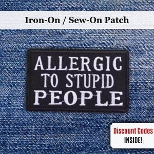 May include: A black, rectangular patch with the white text: "ALLERGIC TO STUPID PEOPLE." The patch is designed to be ironed on or sewn. A small white rectangle with the text "Discount Codes INSIDE!" is also visible.
