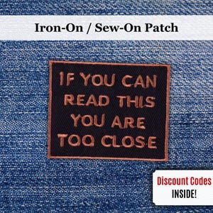 Op de afbeelding: Een zwarte opstrijk- of opnaaibadge met de tekst "IF YOU CAN READ THIS YOU ARE TOO CLOSE" in koperkleurige letters. De badge is rechthoekig en geplaatst tegen een blauwe denim achtergrond. De afbeelding bevat ook een kleine witte coupon met de tekst "Discount Codes INSIDE!"