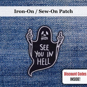 Könnte beinhalten: Schwarz-weißer Aufnäher mit einem Geist, der den Mittelfinger zeigt und den Text "SEE YOU IN HELL" trägt. Der Aufnäher befindet sich auf einem blauen Jeans-Hintergrund.