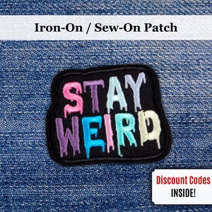 Op de afbeelding: Zwart, rechthoekig opstrijk- of opnaai-patch met de woorden "STAY WEIRD" in kleurrijke, druipende letters. De patch staat op een blauwe denim achtergrond. De patch heeft een zwarte rand.