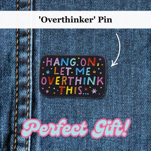 Può includere: Spilla rettangolare nera con la scritta "HANG ON, LET ME OVERTHINK THIS..." in lettere colorate e decorazioni a stella. La spilla è esposta su uno sfondo di denim.
