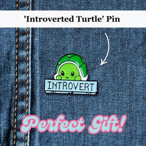 Puede incluir: Un pin de esmalte verde y azul con una tortuga de dibujos animados con la palabra "Introvertido" en una pancarta. El pin está sobre un fondo de mezclilla azul con el texto "¡Regalo perfecto!" en rosa y azul.
