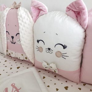 Op de afbeelding: Een set van drie roze en witte wiegbumper met geborduurde dierengezichten. De bumpers hebben een konijn, een kat en een beer. De bumpers zijn versierd met een witte strik en gouden hartjes.
