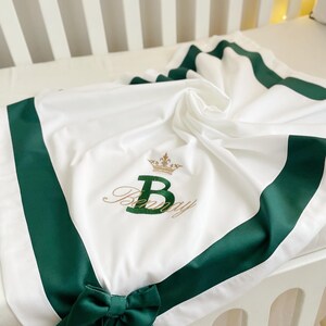 May include: White baby blanket with a green satin border and a green bow. The blanket features an embroidered gold crown and the name "Benny" with a large green "B".