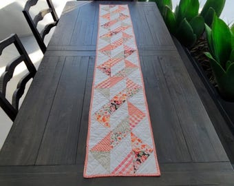 May include: A long, rectangular quilted table runner featuring a repeating geometric pattern of triangles. The design incorporates various fabric prints in shades of orange, coral, and white, finished with a thin orange border and quilted stitching.