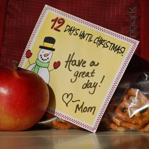 May include: A yellow card with a red stitched border says "12 Days Until Christmas!" and "Have a great day! - Mom". A snowman with a black top hat and a red scarf is in the upper left corner. A red apple is in the lower left corner. A bag of pretzels is in the lower right corner.