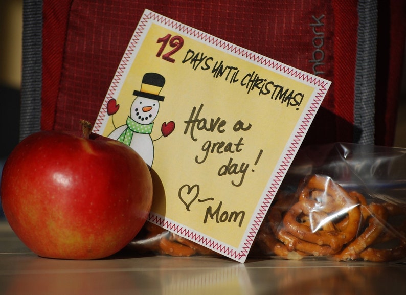 May include: A yellow card with a red stitched border says "12 Days Until Christmas!" and "Have a great day! - Mom". A snowman with a black top hat and a red scarf is in the upper left corner. A red apple is in the lower left corner. A bag of pretzels is in the lower right corner.