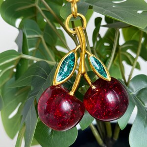 以下が含まれることがあります： 2つの赤いキラキラ光るさくらんぼと緑のキラキラ光る葉が付いた、金色のさくらんぼチャーム。チャームは金のチェーンから吊り下げられています。チャームは緑の葉と白い鉢を背景にしています。