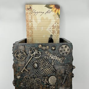 May include: A decorative box with a steampunk design, featuring gears, keys, and a dragonfly. The box holds a notepad with the words "Shopping List" and a sunflower illustration. The box is made of wood and metal.