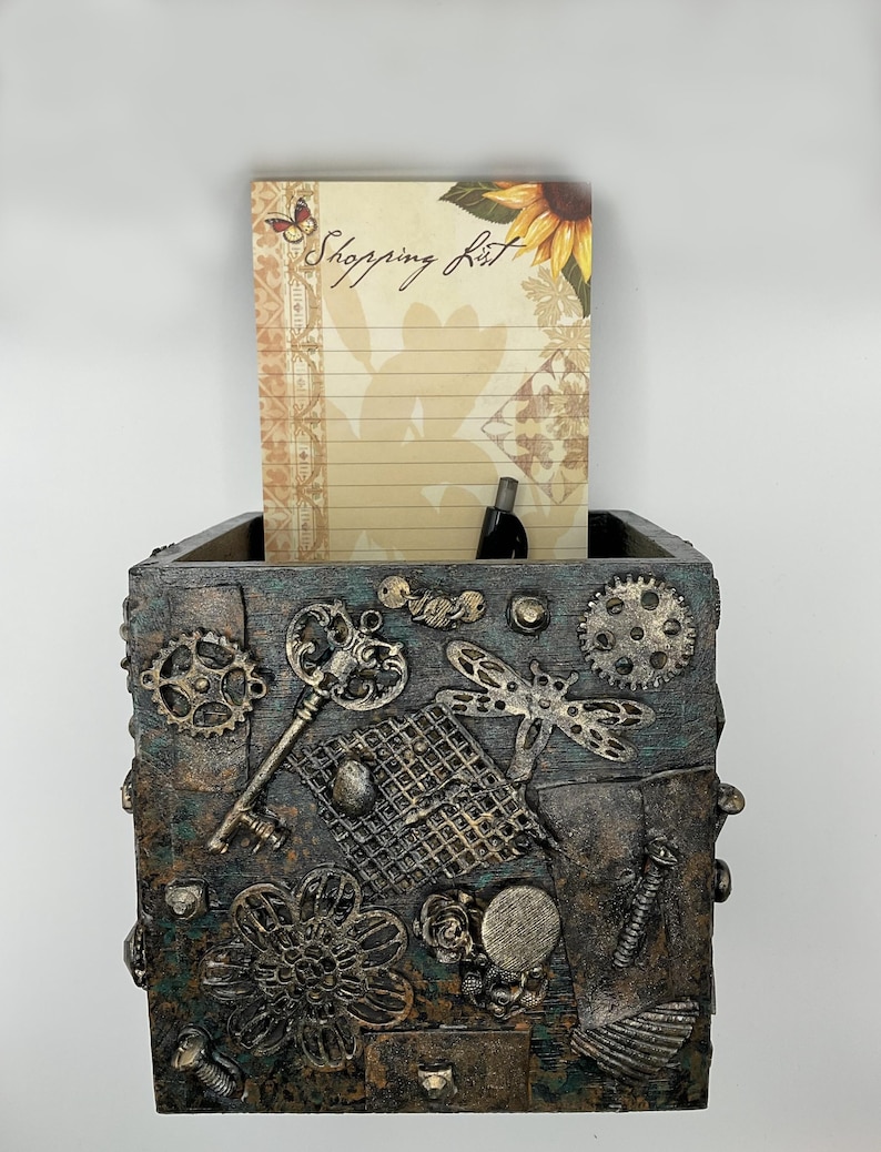 May include: A decorative box with a steampunk design, featuring gears, keys, and a dragonfly. The box holds a notepad with the words "Shopping List" and a sunflower illustration. The box is made of wood and metal.