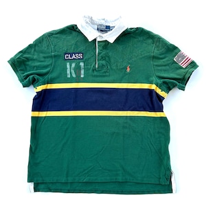 May include: Green polo shirt with a white collar and short sleeves. The shirt features a navy blue and yellow horizontal stripe design across the chest, the text "CLASS K1", and a US flag patch on the sleeve. A small red polo logo is on the chest.