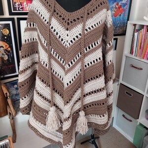 May include: A handmade crochet poncho in shades of brown and cream, displayed on a mannequin. The poncho features a V-neck, chevron pattern, and decorative tassels. The open weave design adds a touch of texture and style to this cozy garment.