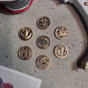 May include: Seven gold-colored Power Rangers coins with different animal symbols on a gray surface. The coins are arranged in a circle with a white Power Ranger helmet and a white Power Ranger sword in the background.