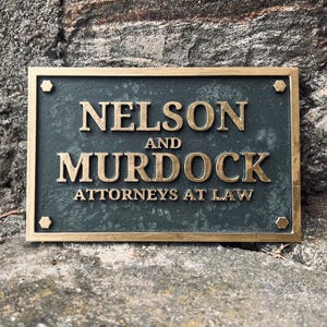 Puede incluir: Una placa de latón con fondo verde oscuro y letras doradas. La placa dice "Nelson and Murdock, Attorneys at Law".