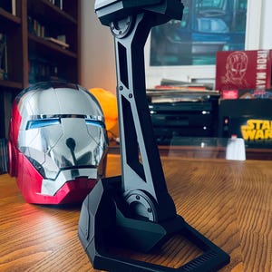 May include: A black headset stand with a geometric design, next to a red and silver Iron Man helmet. The stand has a tall, angular support and a wide base. The helmet has blue eye lights.
