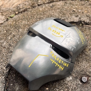 May include: A metallic gray helmet with black accents and yellow text. The helmet has a visor and is marked with the words "TARGETING" and "SENSOR". The helmet appears to be a prop or costume piece.