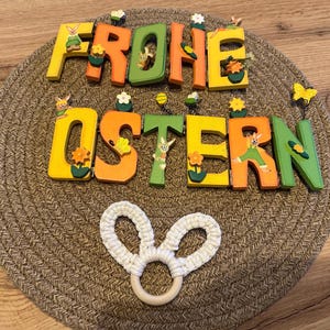 以下が含まれることがあります： ドイツ語で「Frohe Ostern」（ハッピーイースター）と綴られた木製文字。カラフルな花、蝶、ウサギで飾られています。手前に白い編みぐるみのウサギのガラガラがあります。
