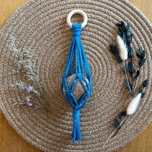 May include: A blue macrame hanging with a round stone inside, hanging from a wooden ring. The macrame is hanging on a brown woven surface.
