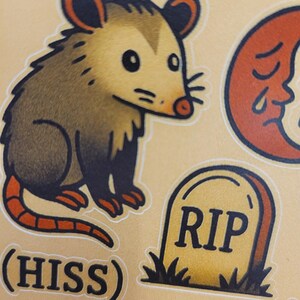May include: A sheet of stickers featuring a cartoon opossum, a crescent moon with a face, a tombstone with "RIP" on it, and the word "(HISS)". The opossum is brown and tan with a red tail.