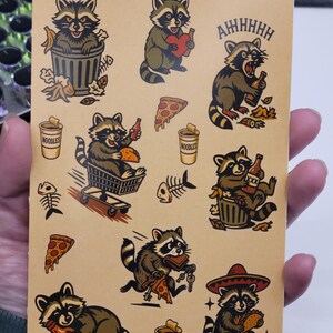 May include: A sheet of raccoon-themed stickers with various illustrations. The stickers feature raccoons with beer bottles, pizza, and trash cans. Some stickers have the word "AHHHHH" and "NOODLES". The stickers are on a tan background.