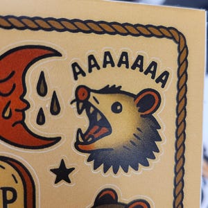 May include: A sticker sheet with a tan background and a brown rope border. The sheet features several illustrations, including a screaming possum with the word "AAAAAAA" above it, a crescent moon, a star, and a tombstone.
