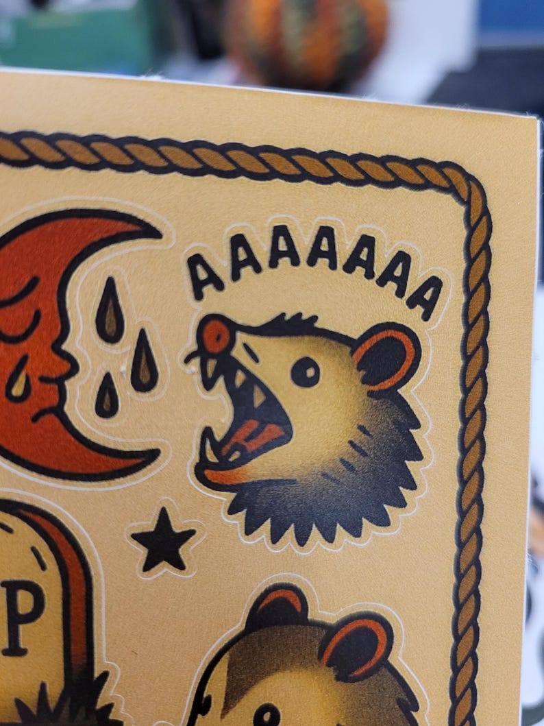May include: A sticker sheet with a tan background and a brown rope border. The sheet features several illustrations, including a screaming possum with the word "AAAAAAA" above it, a crescent moon, a star, and a tombstone.
