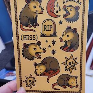 May include: A sheet of stickers featuring opossums in various poses, a crescent moon, a tombstone with "RIP", stars, and skull designs. The stickers are outlined with a brown rope-like border and have the text "(HISS)" and "AAAAAAA".
