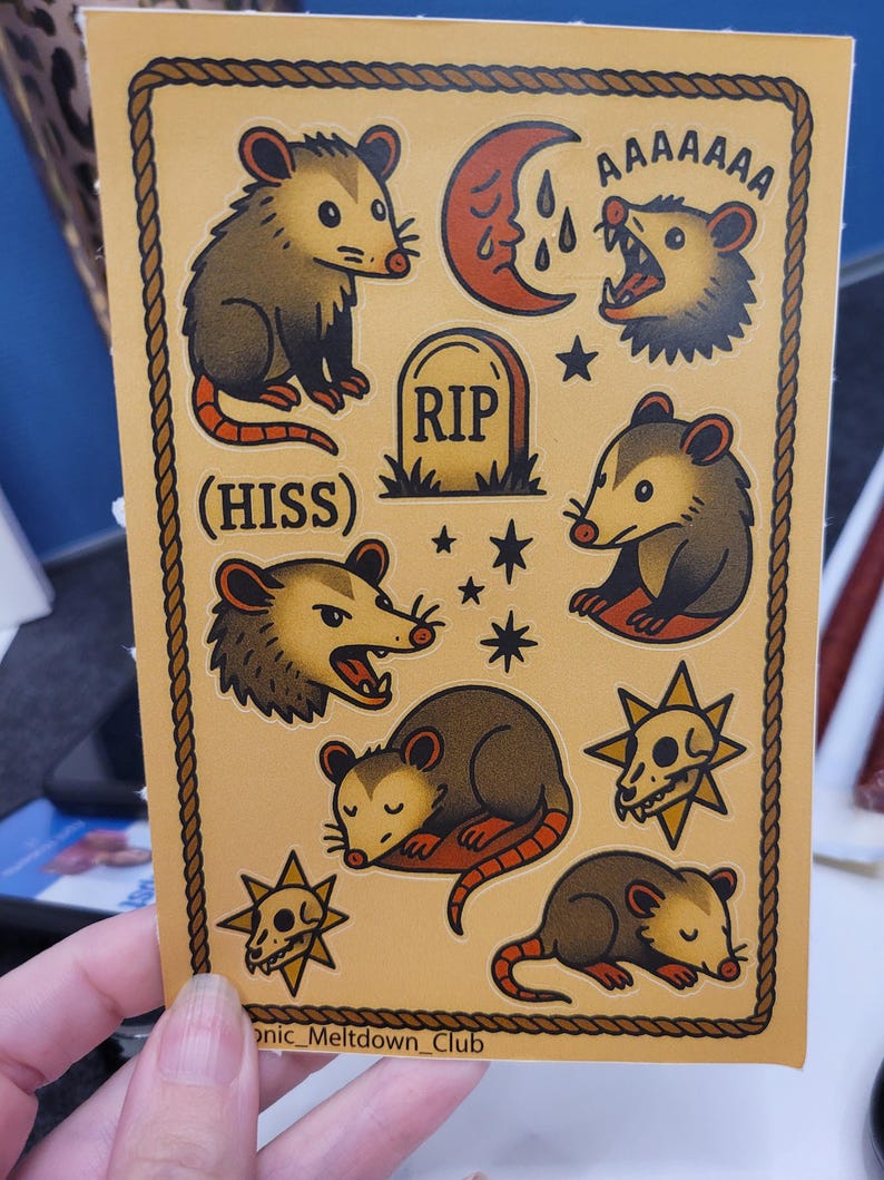 May include: A sheet of stickers featuring opossums in various poses, a crescent moon, a tombstone with "RIP", stars, and skull designs. The stickers are outlined with a brown rope-like border and have the text "(HISS)" and "AAAAAAA".