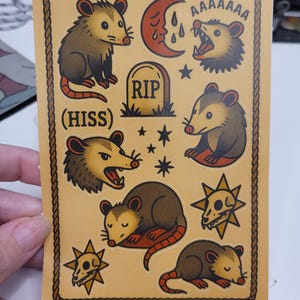 May include: A sticker sheet with a tan background and a brown rope border. The sheet features various opossum illustrations, a tombstone, a crying moon, stars, and a skull. Text includes "(HISS)" and "AAAAAAA". The artist's name is also included.